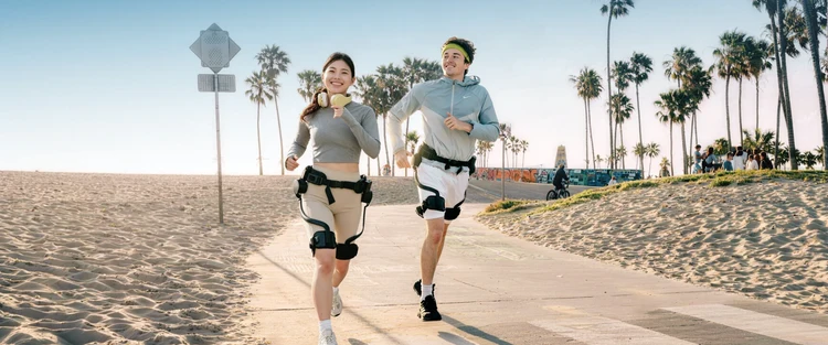 Two people jogging on a beach boardwalk wearing Ascentiz hip-level exoskeletons.