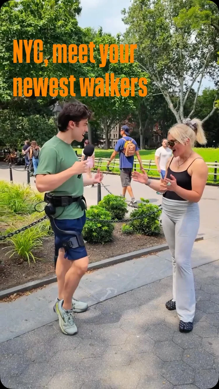 A man wearing Ascentiz H Series exoskeleton stands on a paved path in a park, talking to a woman.