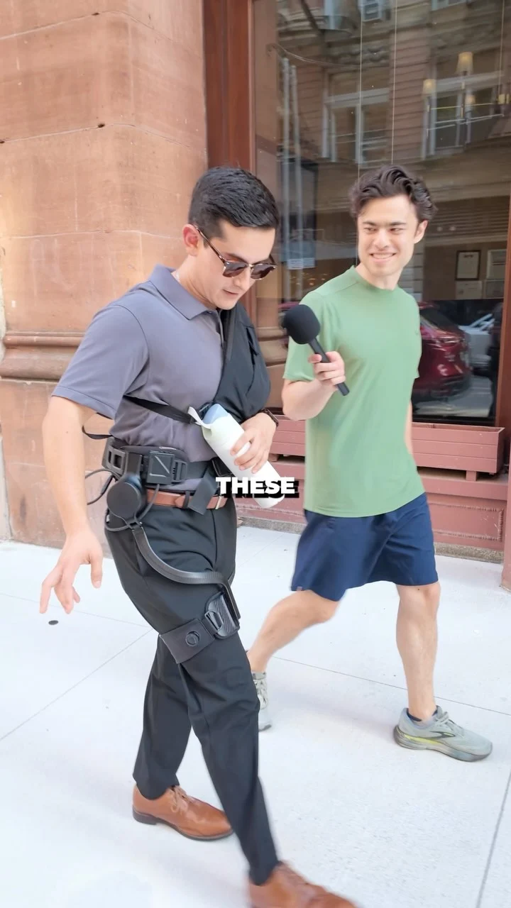 Two men walking outdoors on a city sidewalk. The interviewee is wearing the Ascentiz hip exoskeleton for test.